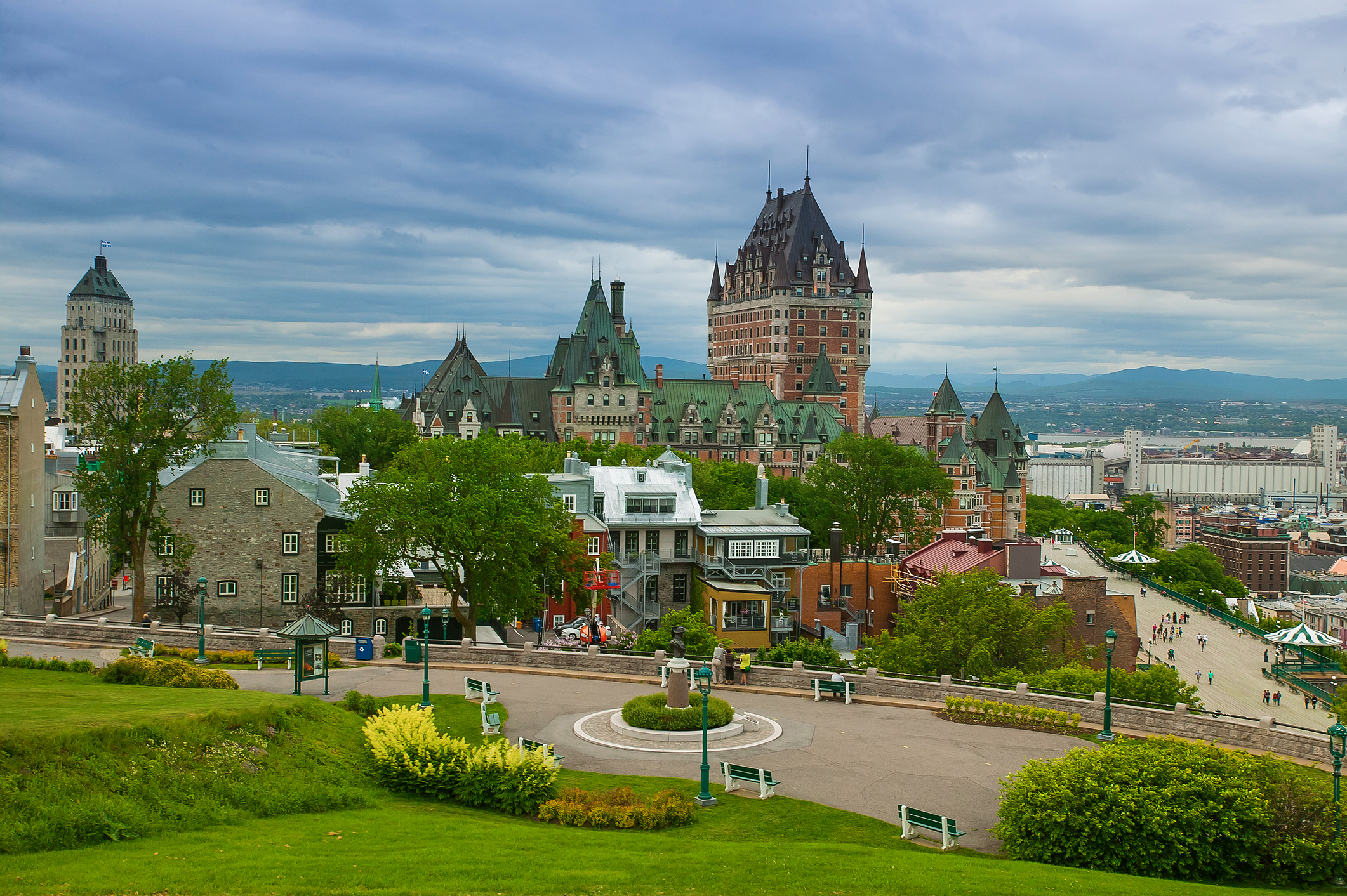The Quebec City skyline from the hill. In front of the buildings there is green gras. The tallest building has a green roof. Ominous skies with grayish clouds.
